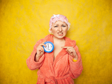 girl in a Terry robe and with a shower cap on her head stands with an alarm clock in her hand and...