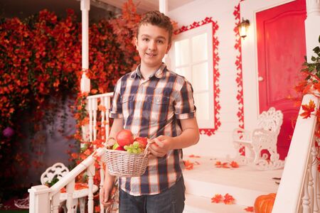 a young man holding a basket of fruit. a teenager in a plaid cozy shirt on the decorated porch of the white house in the fallの写真素材