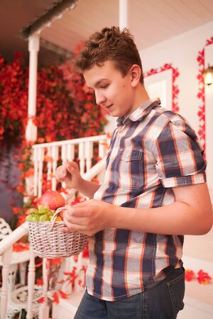 a young man holding a basket of fruit. a teenager in a plaid cozy shirt on the decorated porch of the white house in the fallの写真素材