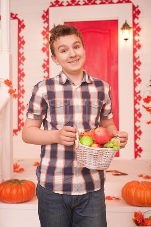 a young man holding a basket of fruit. a teenager in a plaid cozy shirt on the decorated porch of the white house in the fallの写真素材