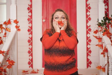 plus size caucasian girl in orange sweater holding autumn red maple leaf in hand with closed face. adult girl stands on the porch of her white house.の写真素材