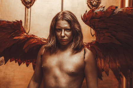 halloween make up. boy ready for party. man is covered in gold paint and poses with large angel wings against the backdrop of large doors in the Studioの写真素材