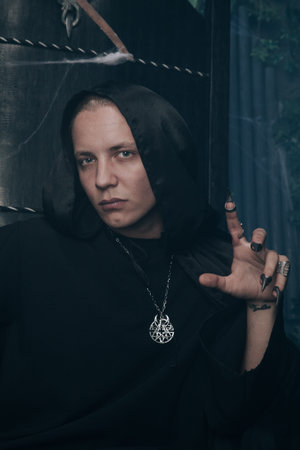 young very angry man with a caucasian face is dressed in a black robe and leather big boots and sits...