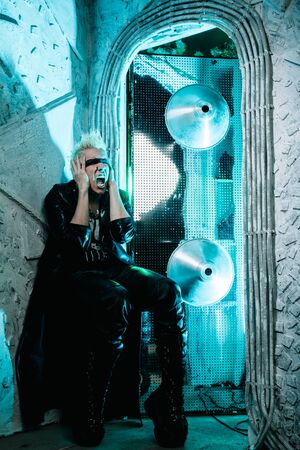 Fashion portrait of the young beautiful alternative man in leather long coat ready for party over silver and neon light studio backgroundの写真素材
