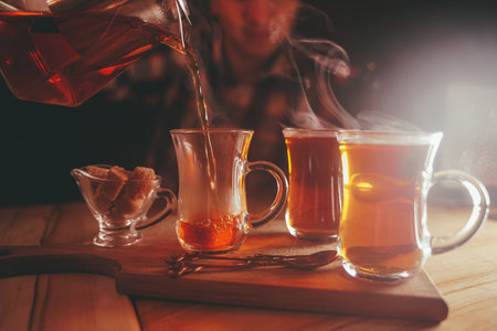 a young man pours tea from a hot teapot into a clear Cup on a wooden table in the evening on dark backgroundの写真素材