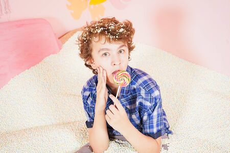 teen boy in the pool with foam soft small white balls. person engaged in relaxation therapy. caucasian kid with colorful lollipop on the stick on pink background.の写真素材
