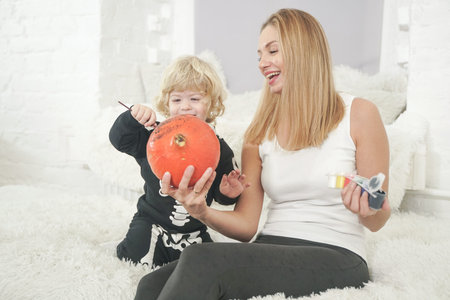 Young woman and her baby son have autumn fun with pumpkin on white windowsill, boy dressed in skeleton costumeの写真素材