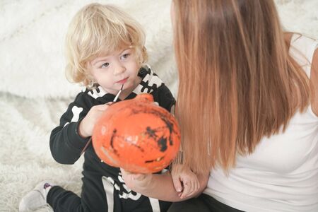 Young woman and her baby son have autumn fun with pumpkin on white windowsill, boy dressed in skeleton costumeの写真素材