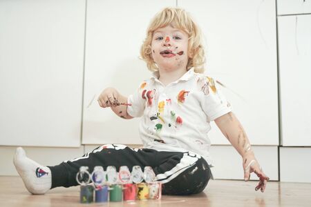 two year old boy with hands and face painted in colorful paints ready for more fun. dirty and happy kid sitting on the floor in white room background.の写真素材