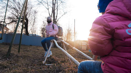 The photo is out of focus, the focus is on the sun. Sunny day, two sisters swinging on a swing near the house, the older sister in a black mask.の写真素材