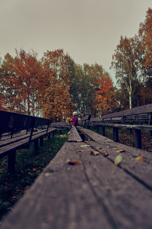 The girl is a child in a crimson cloak in the far term, with a bouquet of yellow leaves, sitting on a bench, bench. The girl sits alone on the bench and looks towards the stage. Selective focus. Spectator seats. autumnの写真素材