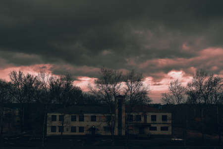 An old building in the village, storm clouds over the roof. Red sunset against the background of trees and buildings. Empty streets and abandoned houses. Eveningの写真素材