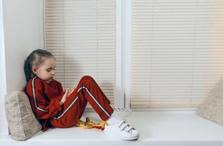 A baby girl in a red suit and pigtails lies on a wide window sill. The window is closed with blinds. Red suit with stripes.の写真素材