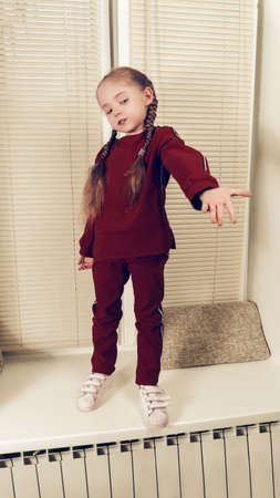 A baby girl with pigtails and a red suit with stripes extends her hand to the side. The girl stands on the windowsill, against the blinds.の写真素材