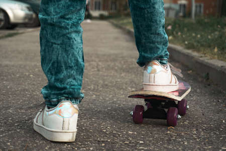 A baby girl stands in a aswalt parking lot and learns to skateboard. Girl in jeans, white T-shirt and white paint with yellow braceletの写真素材