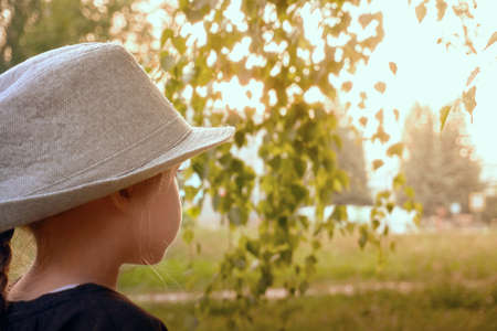 A girl in a black jacket and a gray hat on a sunny summer day looks away, birch branches hang...
