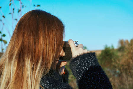A girl in a black coat, holds and photographs with an old film camera silver black. There is a blue sky in the background.の写真素材