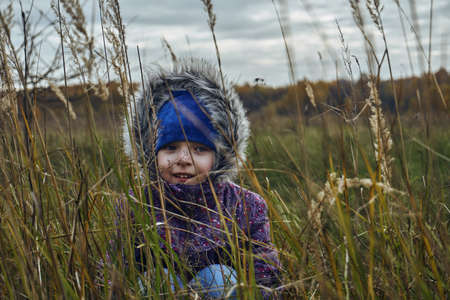 A girl child in a purple jacket and a blue hat sits in the grass and looks straight. Autumn, dry grass and leaves, thick clouds in the sky.の写真素材