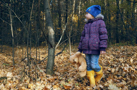 A child in bright rubber yellow boots stands and holds a teddy bear's paw in his hand. Stuffed toy, best friend. Autumn, dry leaves, a walk on a fresh breath.の写真素材
