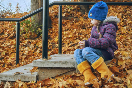 The girl is a child in a blue hat and yellow rubber boots. The girl sits on a concrete step, autumn is a place for outdoor walks.の写真素材