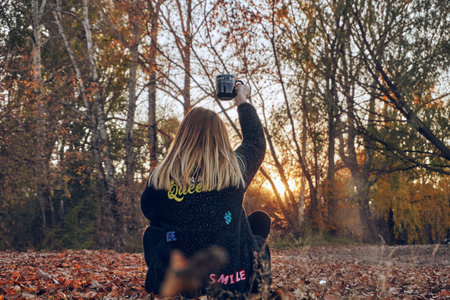 A girl in a black coat sits on dry leaves and holds a black mug with a drink, coffee or tea above...