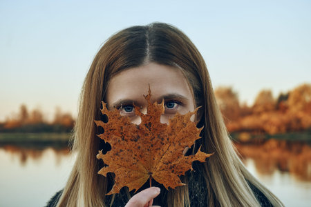 The girl holds a yellow autumn leaf with her hand and looks stubbornly into the camera. Covered...
