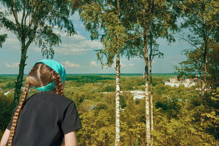 A girl in a scarf looks from a glance or hill at the landscape of nature, white birches stand in...
