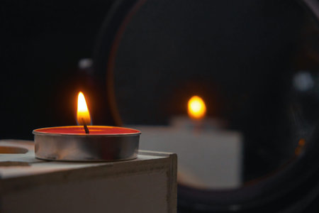 A burning candle stands on a white wooden box, reflection in the mirror and a back black background,...