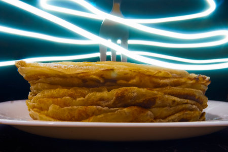 A stack of pancakes on a white plate, a fork stuck in the pancakes, glowing neon lines around the fork. Black background, selective focus.の写真素材