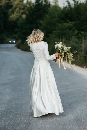 Beautiful elegant bride in lace wedding dress with long full skirt and long sleeves. She is holding a big bouquet of flowers. Outdoors, on the road.