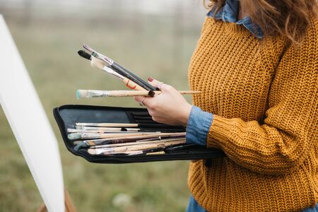 Close up photo of young female artist working on painting outdoors. She holds oil paints, artist brushes, canvas and palette. She is mixing colours on palette. Hands close up.の写真素材