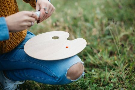 Close up photo of young female artist working on painting outdoors. She holds oil paints, artist brushes, canvas and palette. She is mixing colours on palette. Hands close up.の写真素材