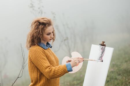 Young female artist working on painting outdoors. She is in front of the canvas and drawing.She holds oil paints, artist brushes, canvas and palette. Portrait of concentrated woman with curly hair.の写真素材