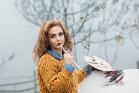 Young female artist working on painting outdoors. She is in front of the canvas and drawing.She holds oil paints, artist brushes, canvas and palette. Portrait of concentrated woman with curly hair.の写真素材