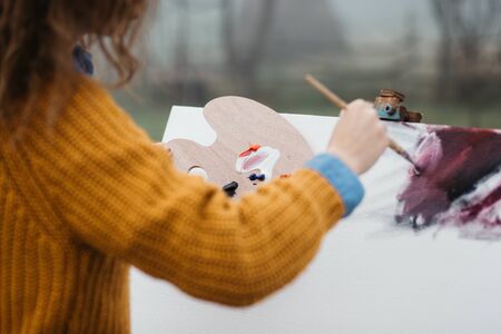Close up photo of young female artist working on painting outdoors. She holds oil paints, artist brushes, canvas and palette. She is mixing colours on palette. Hands close up.の写真素材