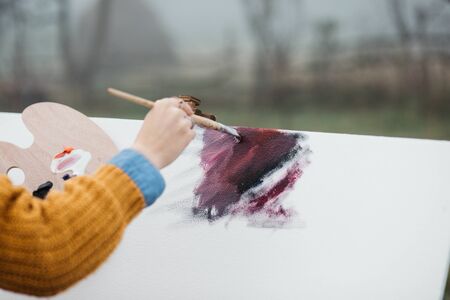 Close up photo of young female artist working on painting outdoors. She holds oil paints, artist brushes, canvas and palette. She is mixing colours on palette. Hands close up.の写真素材
