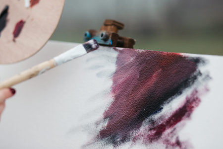Close up photo of young female artist working on painting outdoors. She holds oil paints, artist brushes, canvas and palette. She is mixing colours on palette. Hands close up.の写真素材