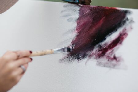 Close up photo of young female artist working on painting outdoors. She holds oil paints, artist brushes, canvas and palette. She is mixing colours on palette. Hands close up.の写真素材