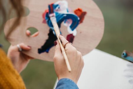 Close up photo of young female artist working on painting outdoors. She holds oil paints, artist brushes, canvas and palette. She is mixing colours on palette. Hands close up.の写真素材
