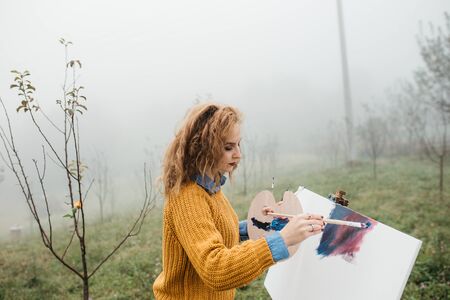 Young female artist working on painting outdoors. She is in front of the canvas and drawing.She holds oil paints, artist brushes, canvas and palette. Portrait of concentrated woman with curly hair.の写真素材