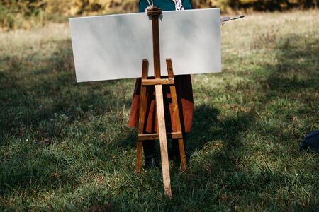 Young female artist working on painting outdoors, in the autumn landscape. She is in front of the canvas and drawing.She holds oil paints, artist brushes, canvas and palette.の写真素材