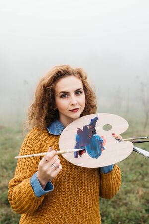 Young female artist working on painting outdoors. She is in front of the canvas and drawing.She holds oil paints, artist brushes, canvas and palette. Portrait of concentrated woman with curly hair.の写真素材