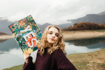 Beautiful woman with curly hair holding painting with sign YES. She is standing on the beach of lake.の写真素材