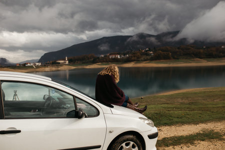 Beautiful woman with curly hair, sitting on the car, holding mobile phone and taking photos of lake...