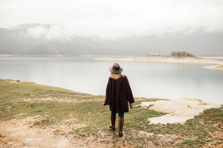 Happy beautiful woman in purple sweater holding her hat and enjoying trip on the lake. Female tourist exploring lake.の写真素材