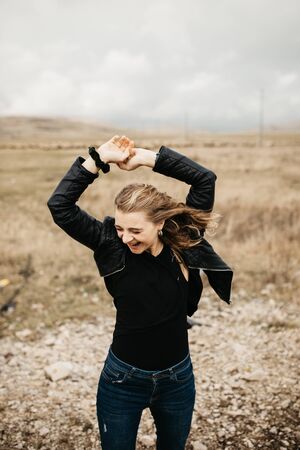 Portrait of young woman with hair blowing in the wind. She is  wearing a leather jacket with a blurred nature and road in background. Wanderlust autumn travel, atmospheric moment.の写真素材