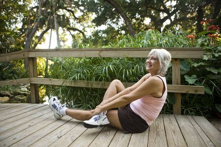 Woman in her 50s in workout clothes and running shoes doing stretching exercises wooden park bridgeの写真素材