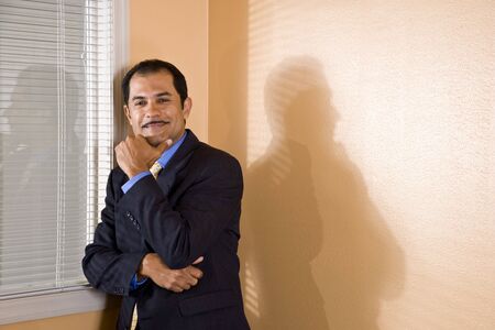 Confident middle-aged Hispanic businessman standing by office window with hand on chinの写真素材