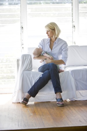 Mother holding sleeping 3 month old baby on sofa in sunny room at homeの写真素材