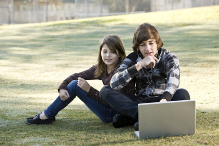 Boy and girl sitting on grass with laptop, online in parkの写真素材